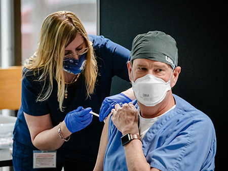 Dr. David Pigott, MD (Professor, Emergency Medicine) is wearing blue scrubs and a PPE (Personal Protective Equipment) head cover, and face mask while receiving the COVID-19 (Coronavirus Disease) vaccination from a female healthcare worker wearing scrubs and a PPE face mask and gloves, December 2020.