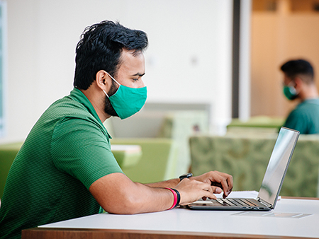 From side, a male student, Dhruv Singh (Student, Mechanical Engineering), is wearing a face mask and studying on a laptop computer while sitting at a table in the lobby of the Collat School of Business during the COVID-19 (Coronavirus Disease) pandemic while another male student is wearing a face mask and sitting in the background, September 2020.  