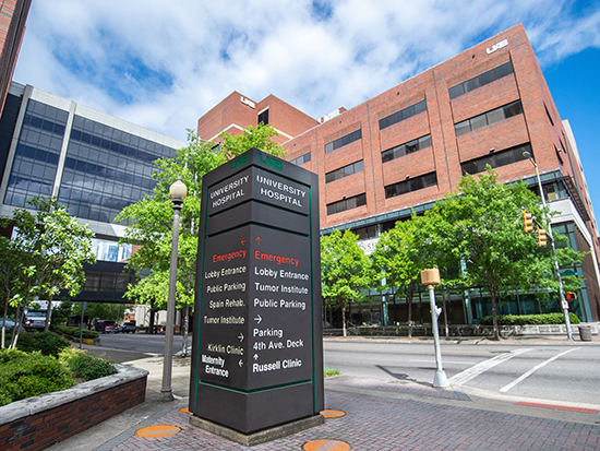 UAB University Hospital signage on sidewalk with the O'Neal Comprehensive Cancer Center, the North Pavilion, and blue sky and white clouds in the background, April 2020.