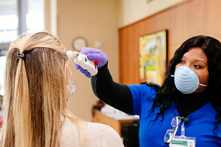 Health care worker is wearing a PPE (Personal Protective Equipment) face mask and gloves while taking the temperature of a female patient. 