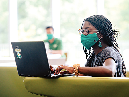 From side, a black female student, Kafui Owusua Sakyi-Addo (Student, Pscychology), is wearing a UAB-branded face mask and studying on a laptop computer while sitting in a chair in the lobby of the Collat School of Business during the COVID-19 (Coronavirus Disease) pandemic while a male student is sitting in the background, September 2020.