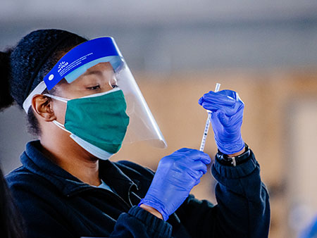COVID-19 (Coronavirus Disease) Vaccination Site in UAB-Highlands parking deck. From side, black female health care worker is wearing PPE (Personal Protective Equipment) gloves, face mask, and face shield while administering vaccine into a syringe on January 19, 2021. 