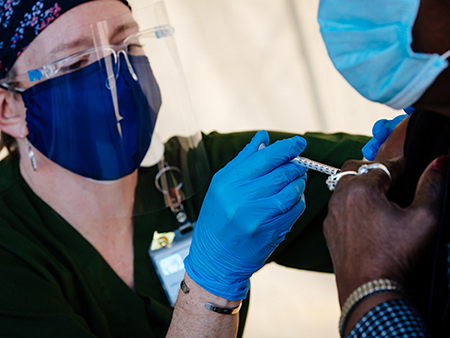 A female healthcare worker is wearing PPE (Personal Protective Equipment) head cover, face mask, face shield, and gloves while administering the COVID-19 vaccine to a black male at the UAB COVID-19 Vaccination Site at Parker High School on February 10, 2021/.