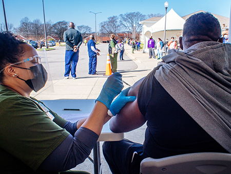 From side, a Black female healthcare worker is wearing PPE (Personal Protective Equipment) face mask and gloves as she vaccinates a Black male at the UAB COVID-19 Vaccination Site at Parker High School. 