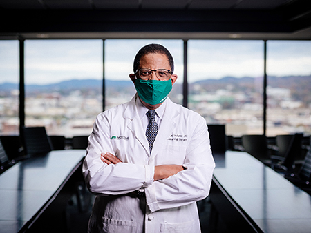 Dr. Selwyn Vickers, MD (Senior VP/Dean, School of Medicine) is wearing white medical coat and face mask due to the COVID-19 (Coronavirus Disease) pandemic while standing beside table in conference room, December 2020.