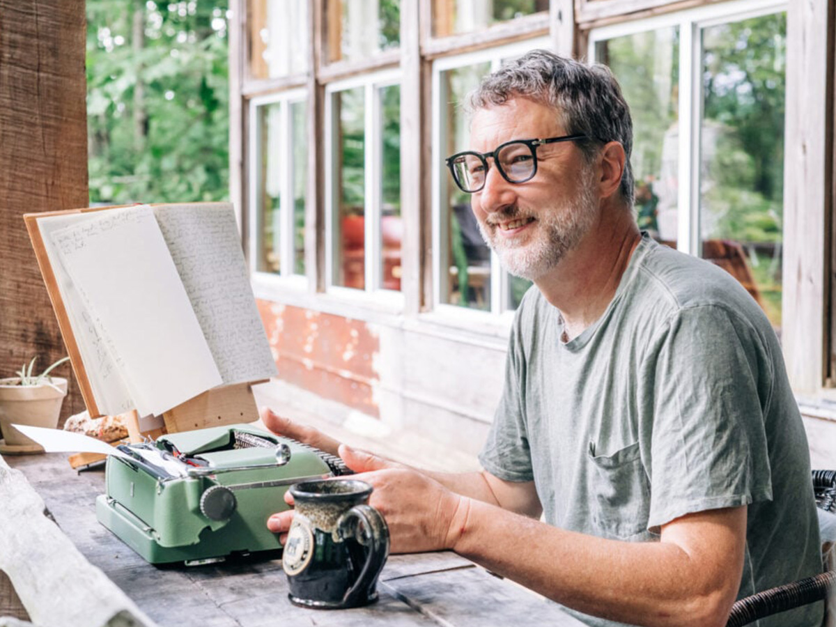 A man smiles while seated outdoors on a deck, typing on a small typewriter. 