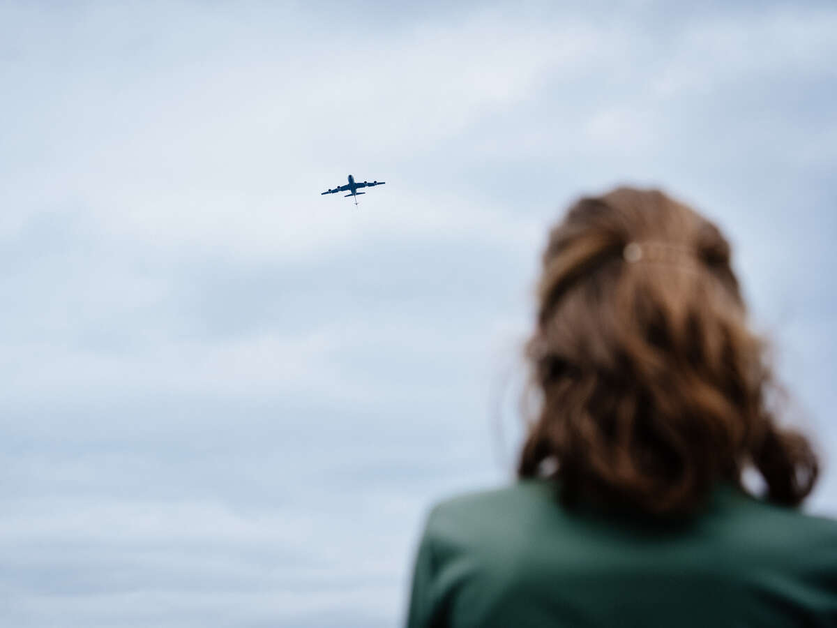 Back, a female health care worker, wearing green scrubs, is looking at the sky as the airplane flies.
