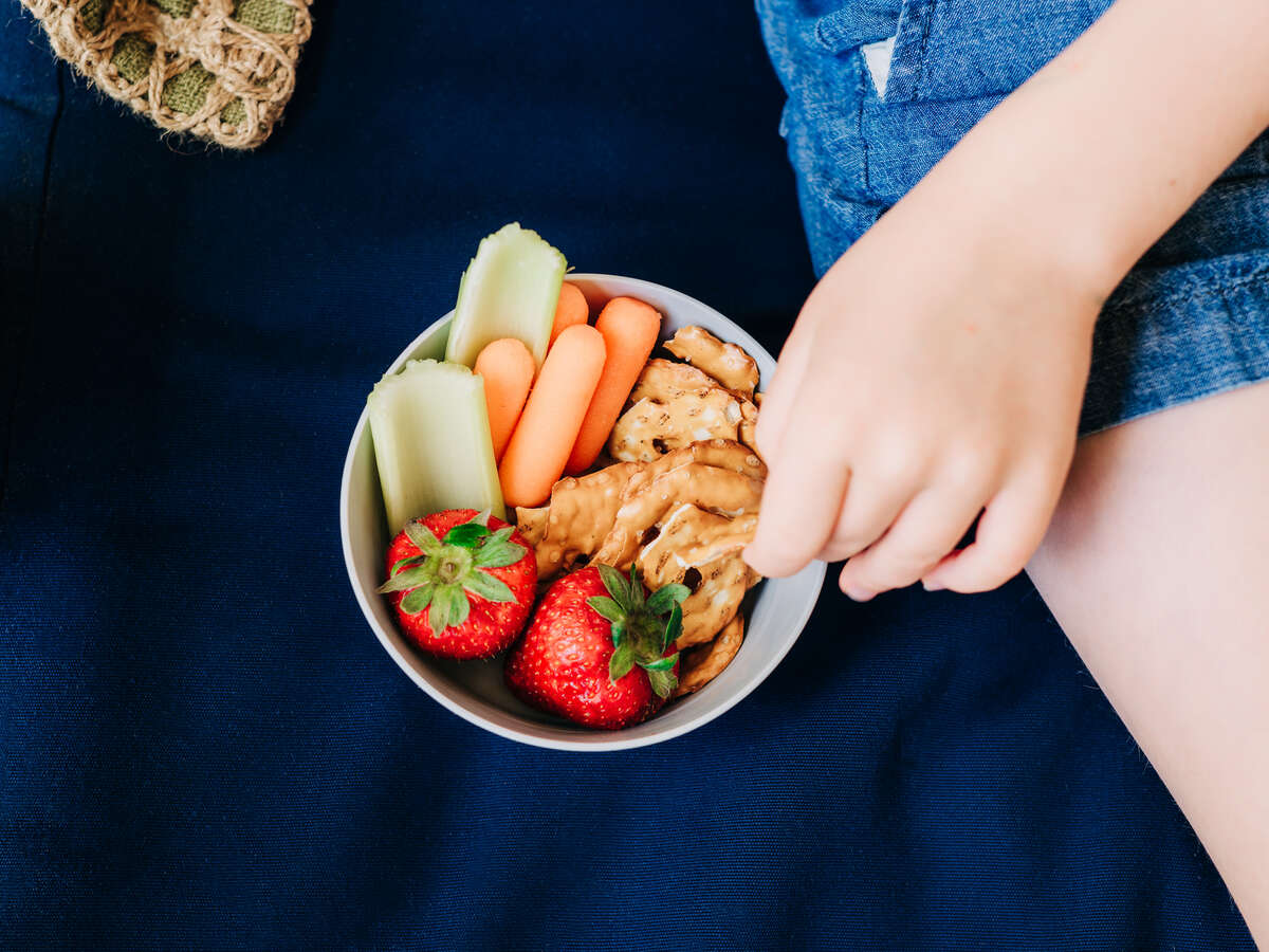 Shot from above, close-up of boy's hand is reaching into a bowl of fresh strawberries, baby carrot, celery pieces, and pretzels