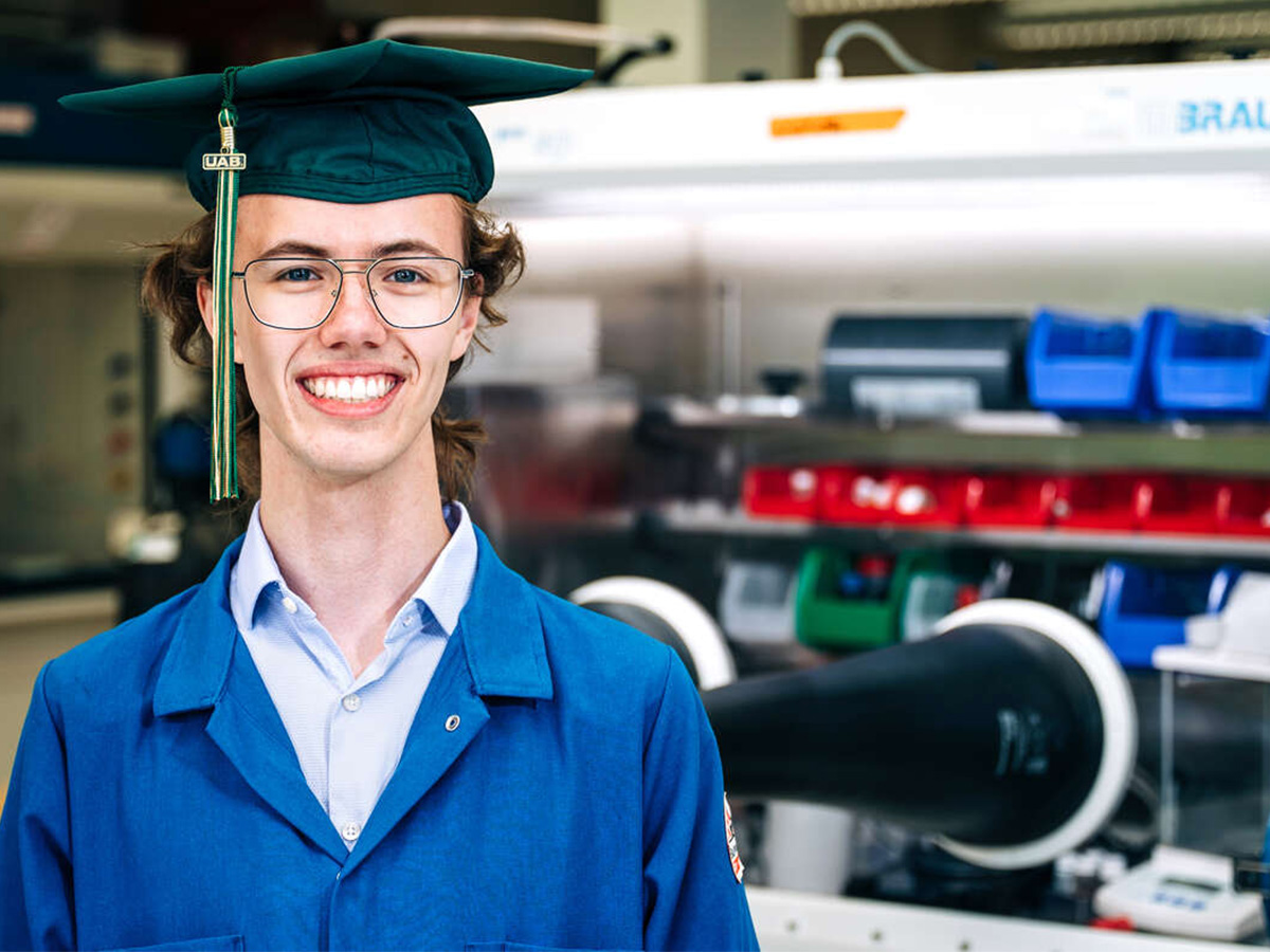Image of Mason Bayles, UAB graduate, in green graduation cap and blue lab coat standing in from of lab equipment. 