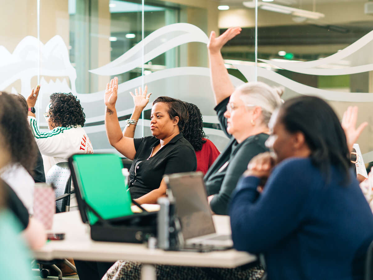 A room full of people sitting at desks raise their hands in an interactive moment of teaching.