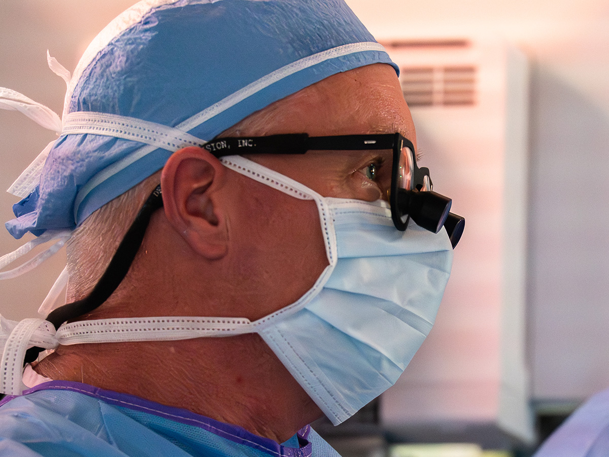 A close-up profile of surgeon Michael Hanaway, M.D., wearing a blue surgical cap, a face mask, and specialized magnifying loupes attached to his glasses.