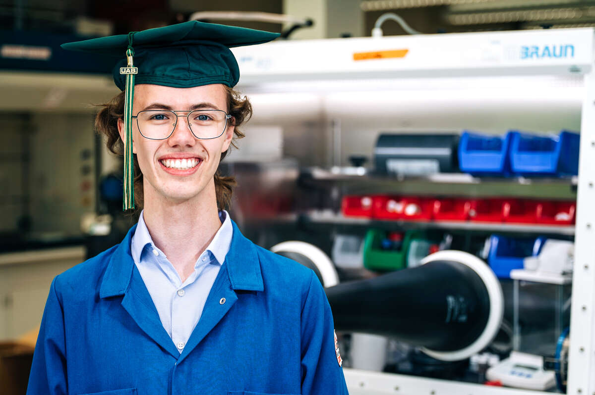 Image of Mason Bayles, UAB graduate, in green graduation cap and blue lab coat standing in from of lab equipment. 