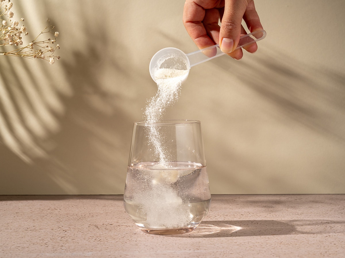 Image of a hand dumping a scoop of collagen powder into a clear glass of water. 