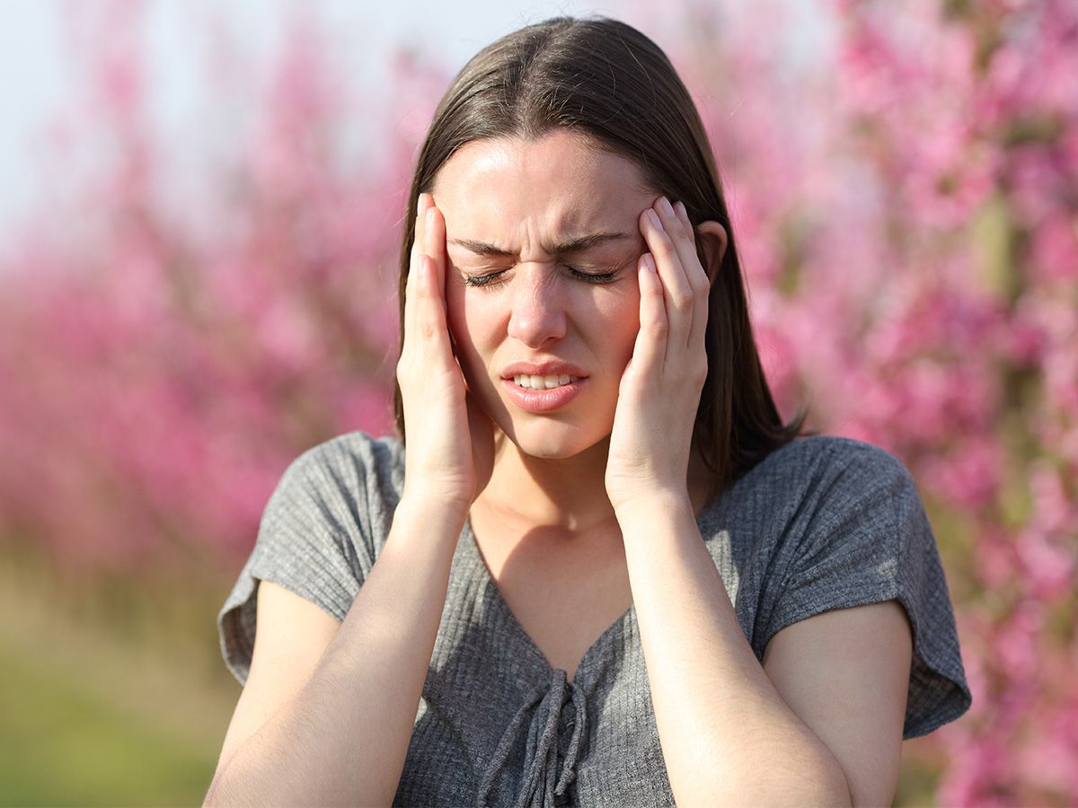 Person standing outdoors with eyes closed, pressing both hands to the sides of the head, with pink flowering trees blurred in the background.
