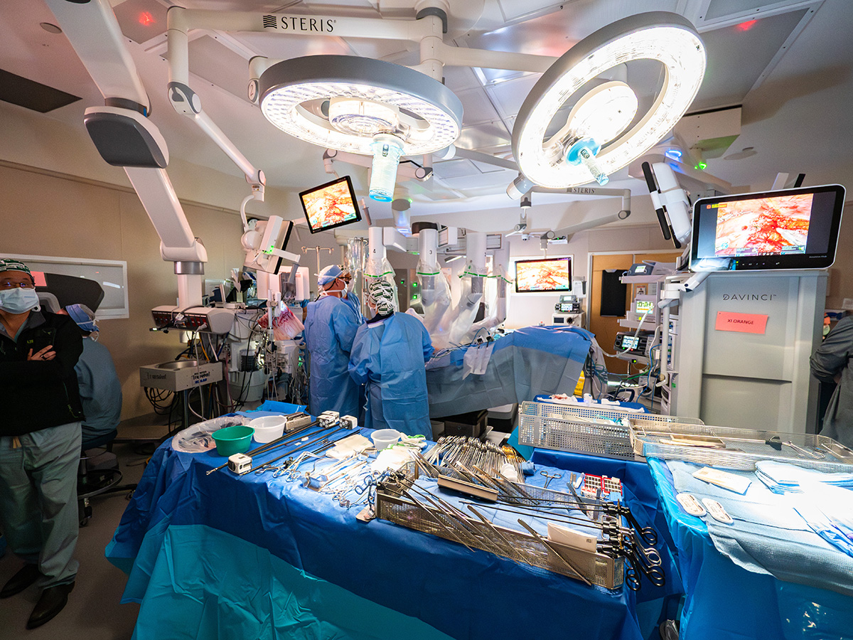 A wide view of a high-tech operating room at UAB Hospital featuring a surgical team, robotic surgery system, multiple monitors displaying surgical footage, and a table of sterile instruments.