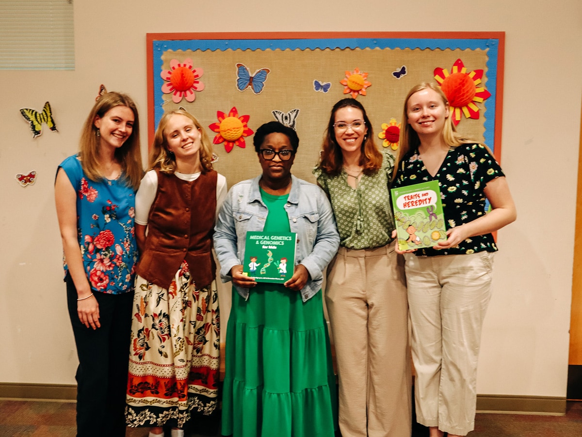 Group photo of UAB genetic counseling students holding books in front of a colorful library background. 