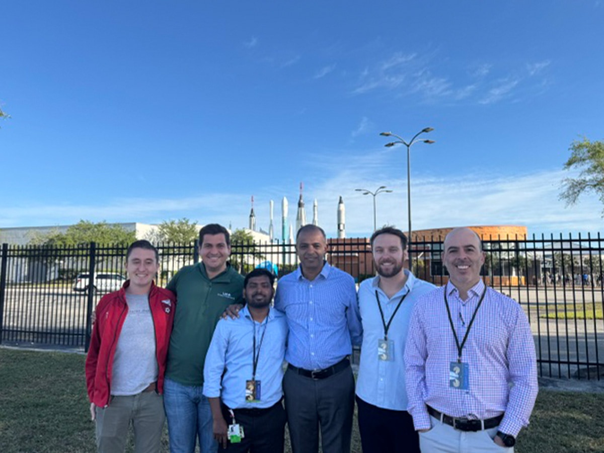 Outside image of researchers standing in front of fence with space rockets in the distance. 