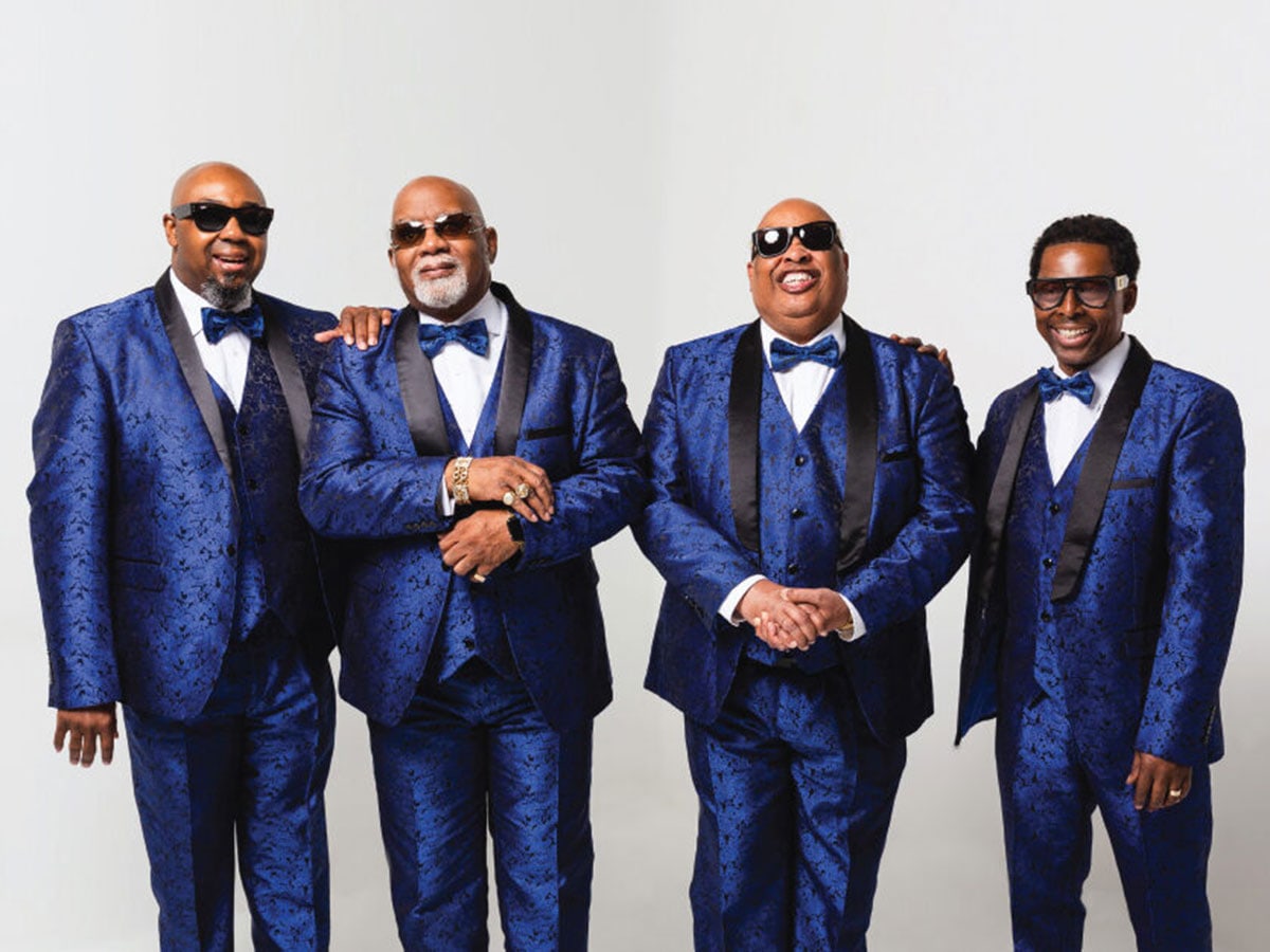Four men wearing glasses and matching royal blue suits and bowties stand together and smile. 