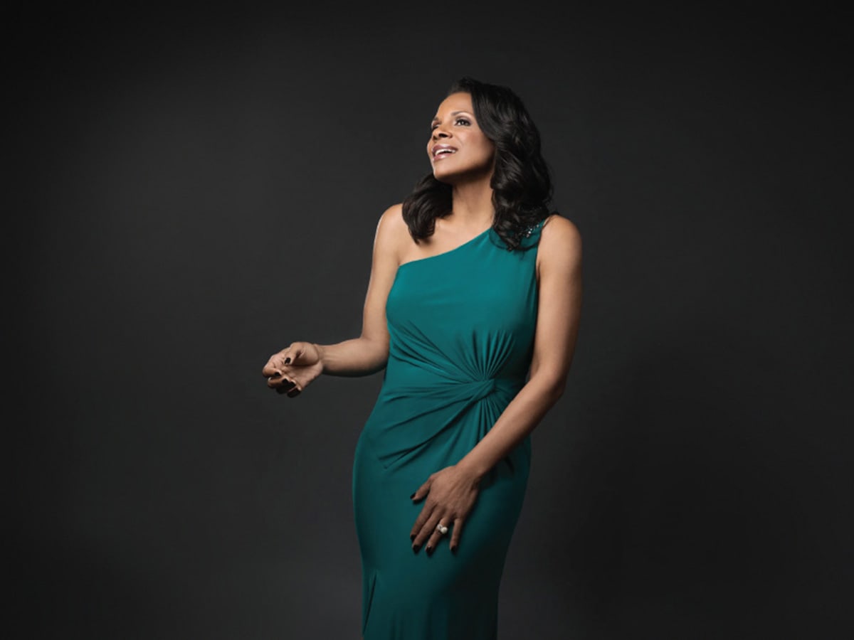 A woman in a green dress poses in front of a dark background. 