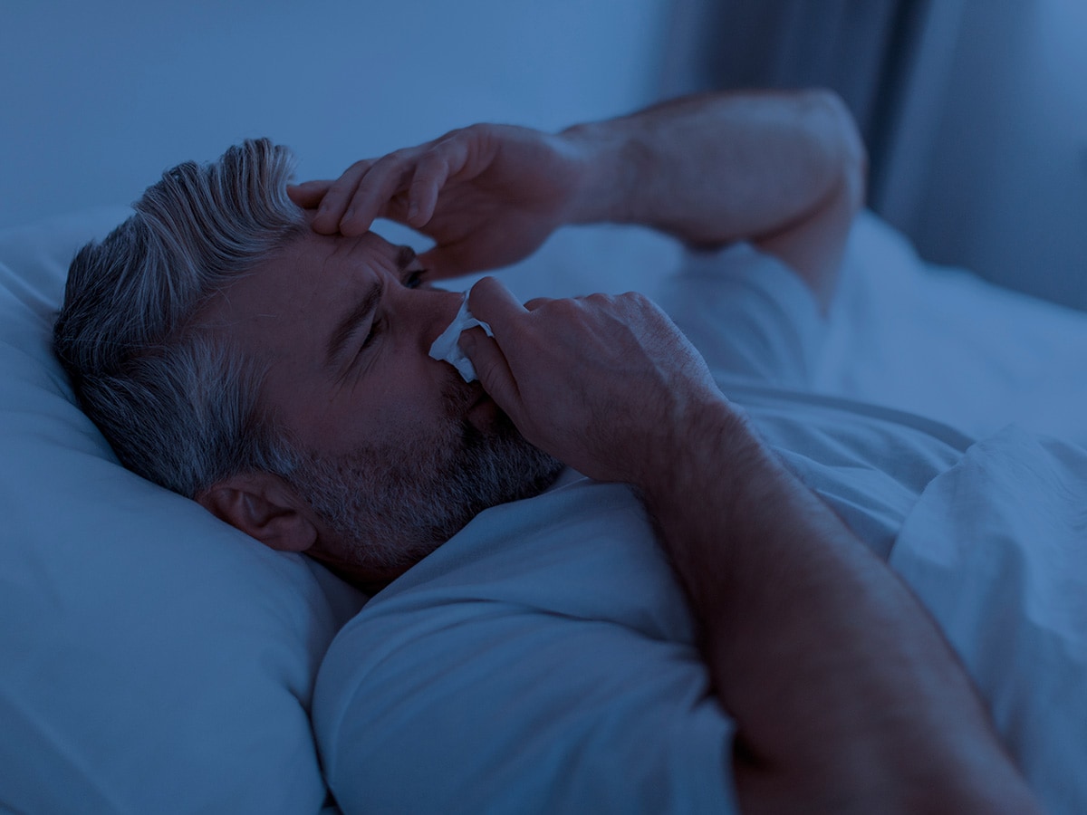 Man lies in bed with a tissue covering his nose with his right hand and his left hand on forhead.