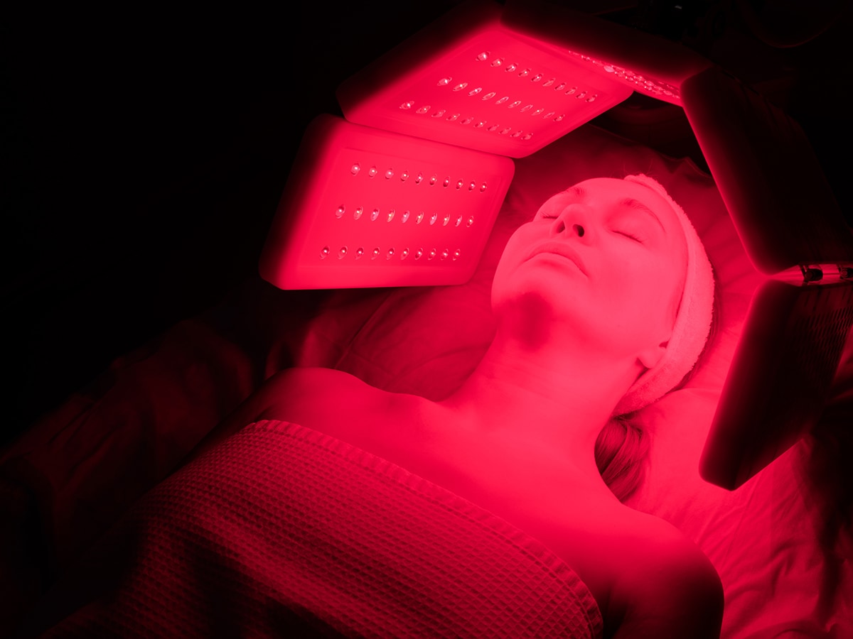 Woman using red light therapy device aimed at the face while lying down.