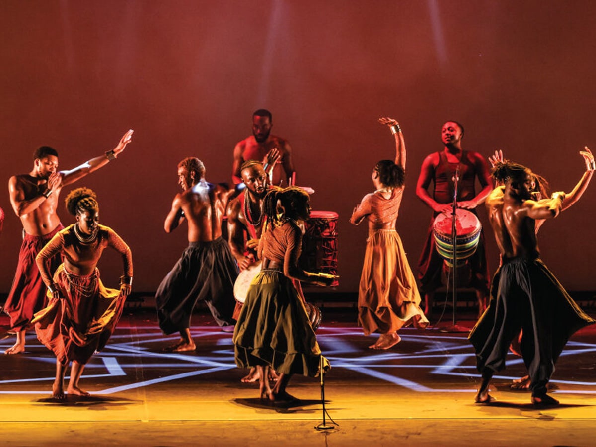 Performers dressed in billowing pants and skirts dance barefoot on stage and play drums during a performance.