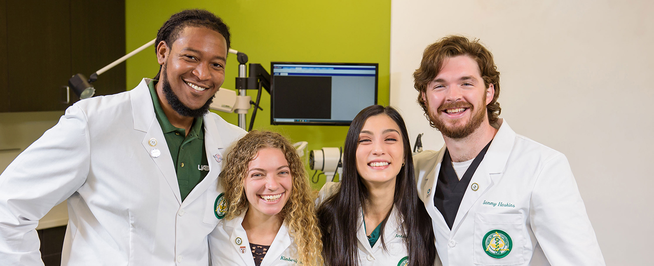 Four dentistry studentds, two male, two female, posing with arms around each other inside the school and smiling.