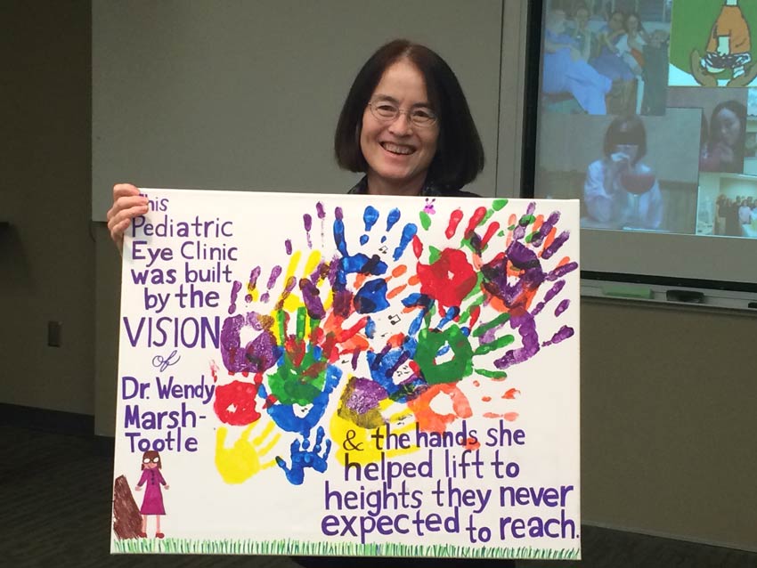 Dr. Wendy Marsh-Tootle smiling and holding a painted sign decorated with handprints which reads "The Pediatric Eye Clinic was built by the vision of Dr. Wendy Marsh-Tootle and the hands she helped lift to heights they never expected to reach".