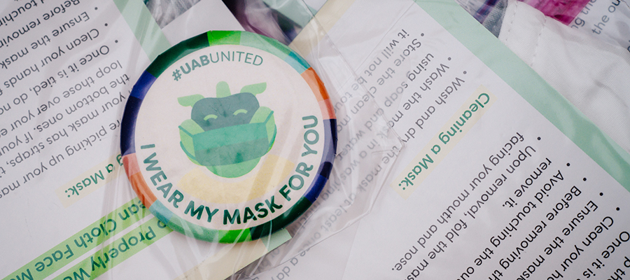 Close-up of a button that reads "#UABUNITED - I WEAR MY MASK FOR YOU" over a stack of instructions for how to wear and clean cloth face masks at a mask distribution drive-thru in Express Lot No. 4 during the COVID-19 (Coronavirus Disease) pandemic, August 2020.