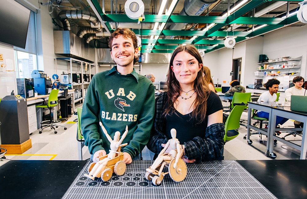 Tony Tamburello and Cam Boydstun pose with small human figure in a wheelchair in the Design x Prototyping Lab