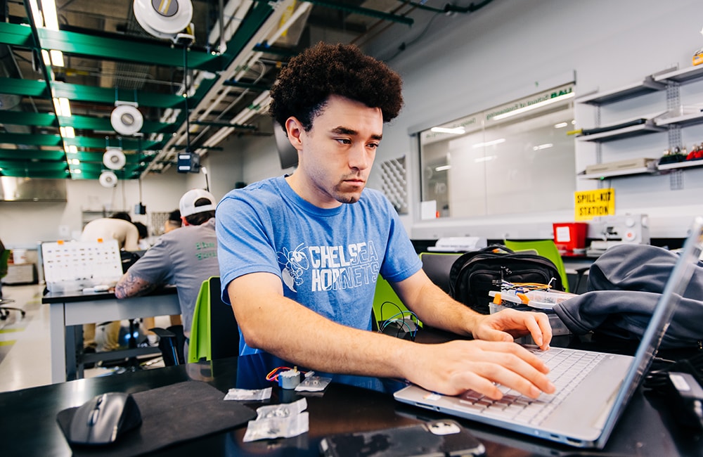 Alex Johnson sits at a table in the Design x Prototyping Lab at the UAB School of Engineering, working on his laptop to develop his Braille teleprompter device prototype