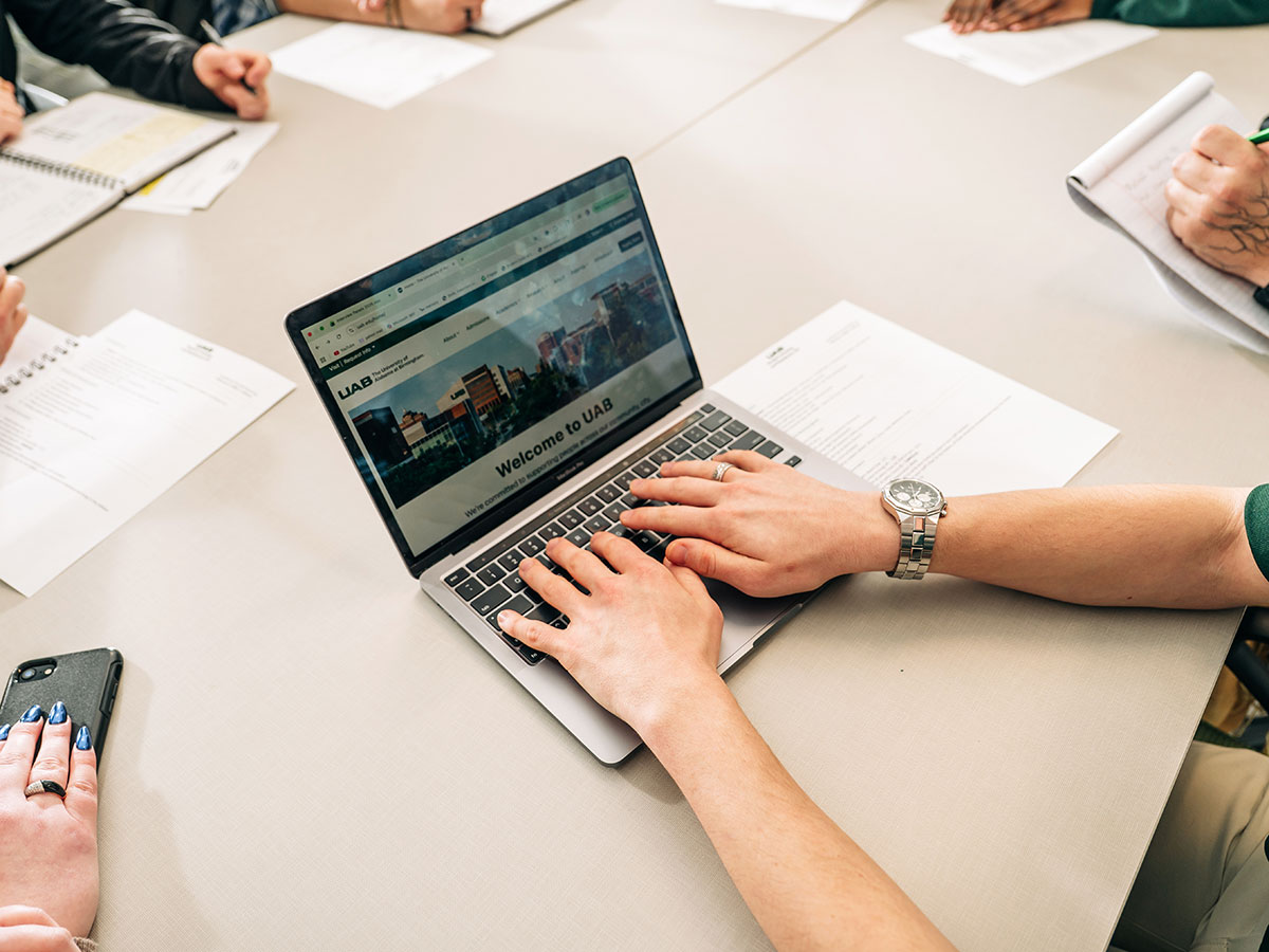 Closeup of an employee's hands on a laptop open to the UAB website in a staged staff meeting on campus