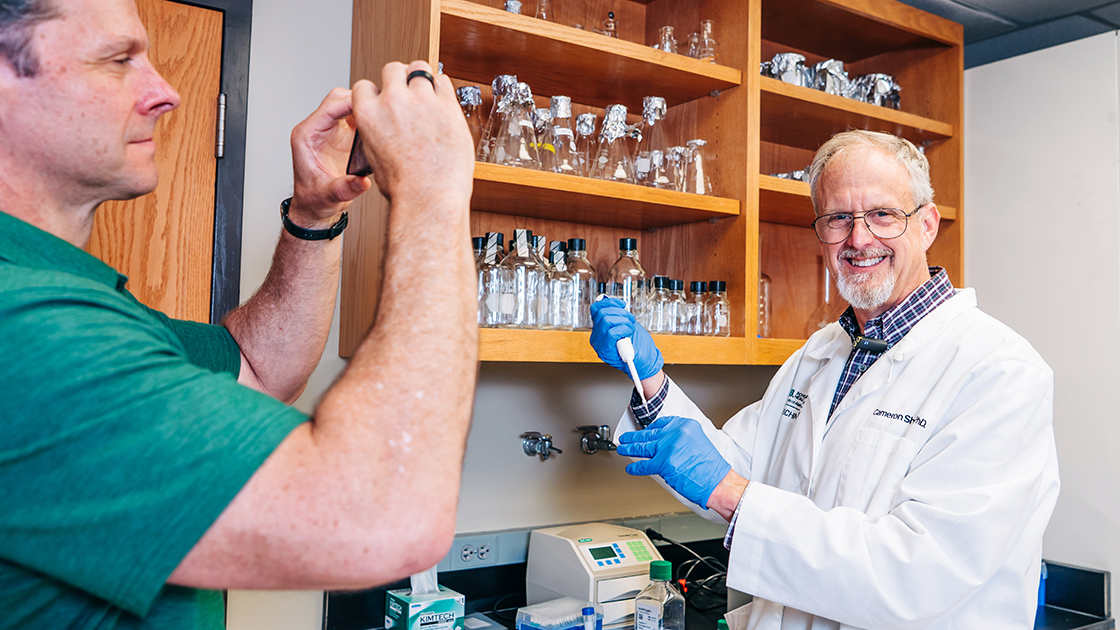 Cameron Sheeler, Ph.D., looks at the camera while pipetting and Kevin Storr holds his phone filming