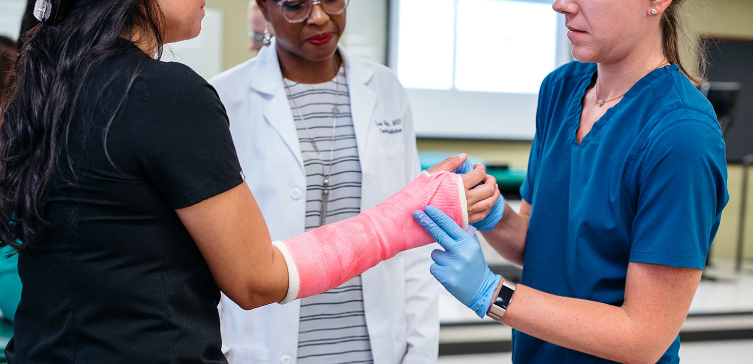 Person in scrubs touching patient's arm that is in a cast while person in a lab coat looks on.
