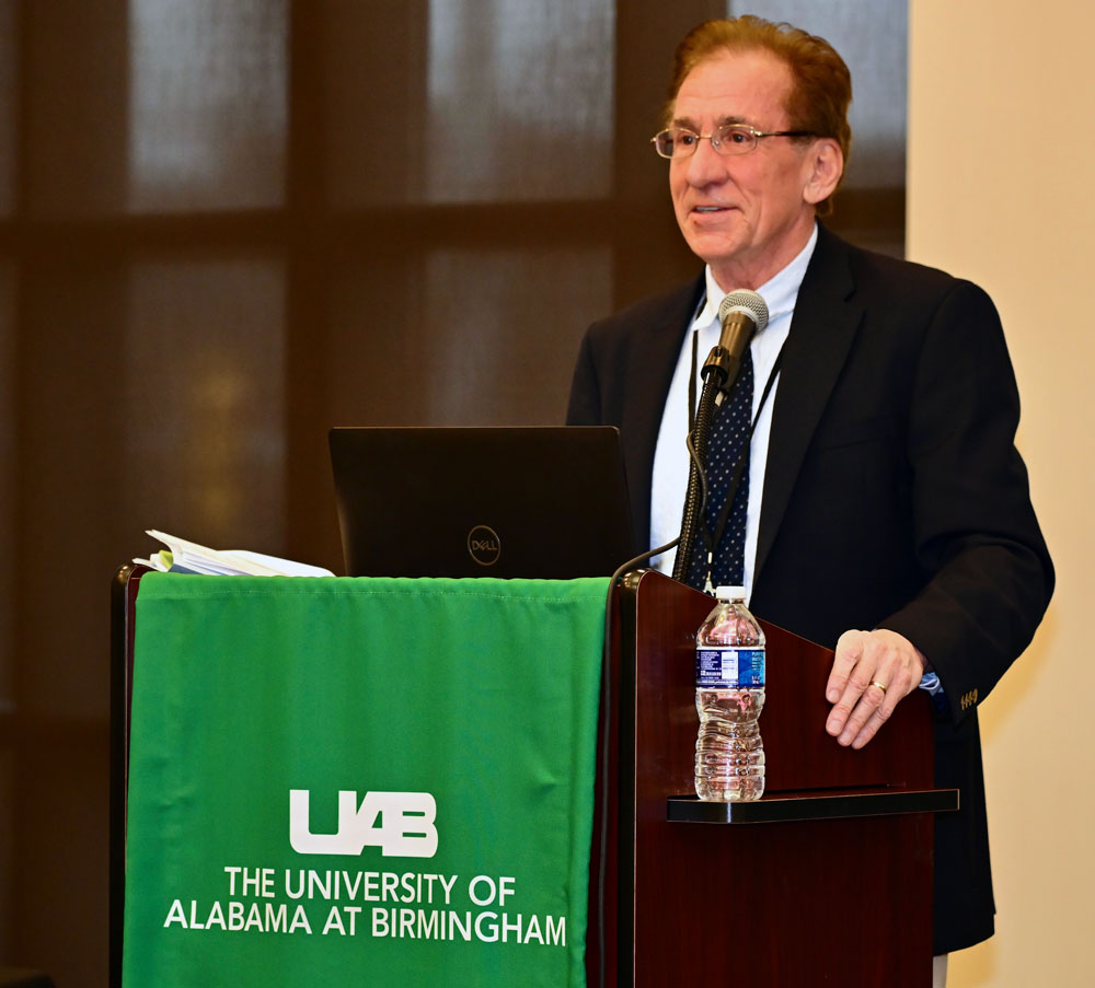 Dr. Rimmer at a podium with a green UAB banner addressing the symposium. 