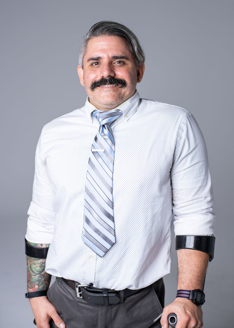 Dr. Mendonca has short salt-and-pepper hair and a large dark mustache. He wears a light blue button-up shirt with a short, wide light-blue striped tie. A tattoo sleeve is visible on one wrist, and the tops of his assistive devices are visible. 