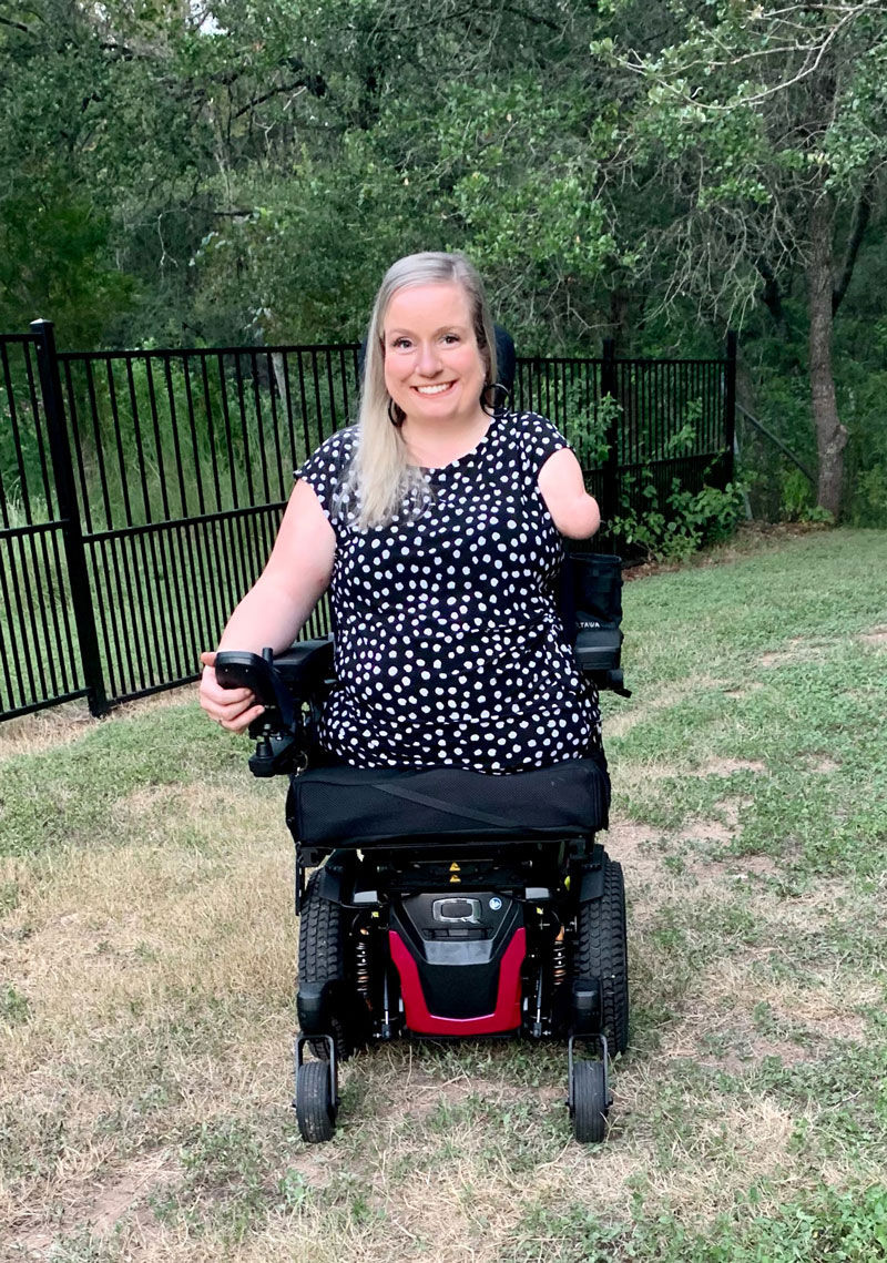  Dr. Andrews, outside in a green space in her powered chair. She has long blond hair and is wearing a black blouse with white polkadots. 