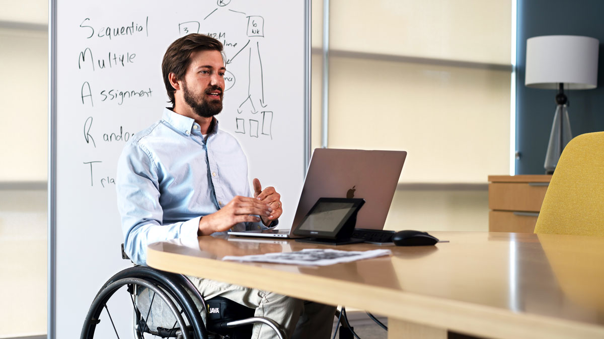 Bearded man sits in wheelchair at a desk with a laptop, a whiteboard behind him.