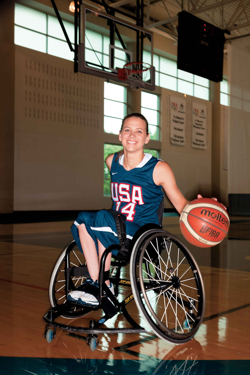 Cook dribbling a basketball in her chair wearing her USA 14 jersey, on a basketball court. 