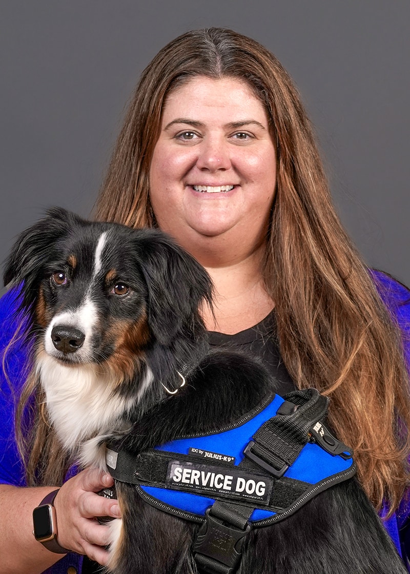 Mills holds her service dog, a minature Australian shepherd, which is wearing a blue service vest.