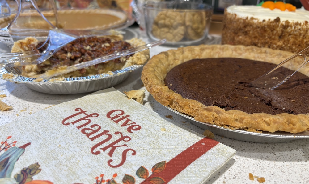 Thanksgiving desserts pecan pie, chocolate pie, pumpkin pie, carrot cake and cookies. The napkin in the foreground has autumn design and says "Give Thanks"