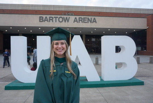 Lizzy Bumpas dressed in UAB graduation cap and gown in front of giant UAB letters at Bartow Arena.