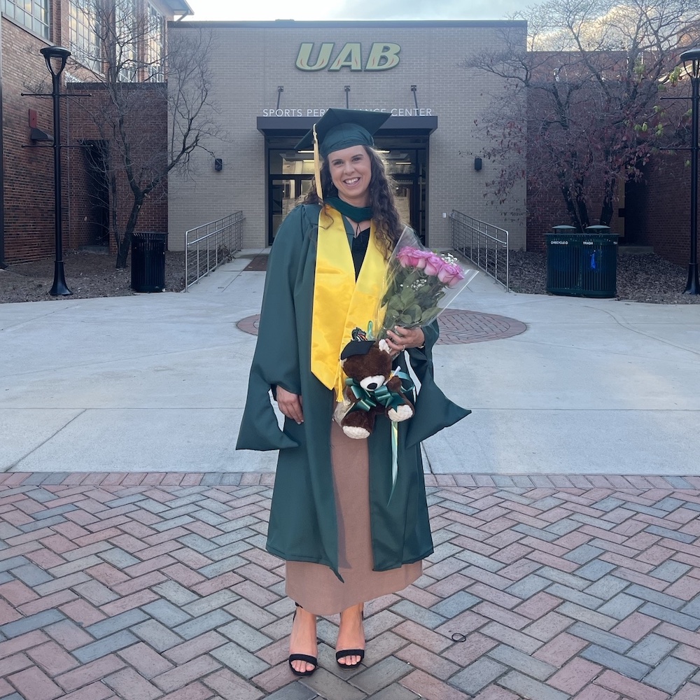 Danielle North in UAB graduation cap and gown standing in front of the Sports Performance Center.