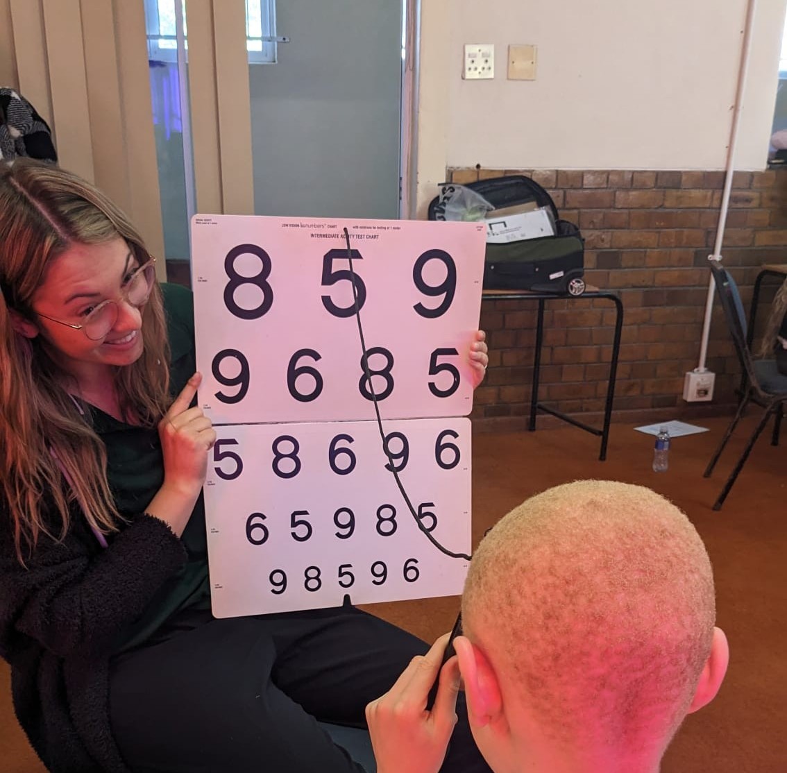 Smiling UAB OT student holds a numbered eye chart for a South African child to read.