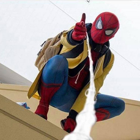 Er in a costume from Spider-Man: Homecoming, in spider suit, yellow high-school jacket, and backback, crouched on a wall and shooting webs at the camera. 