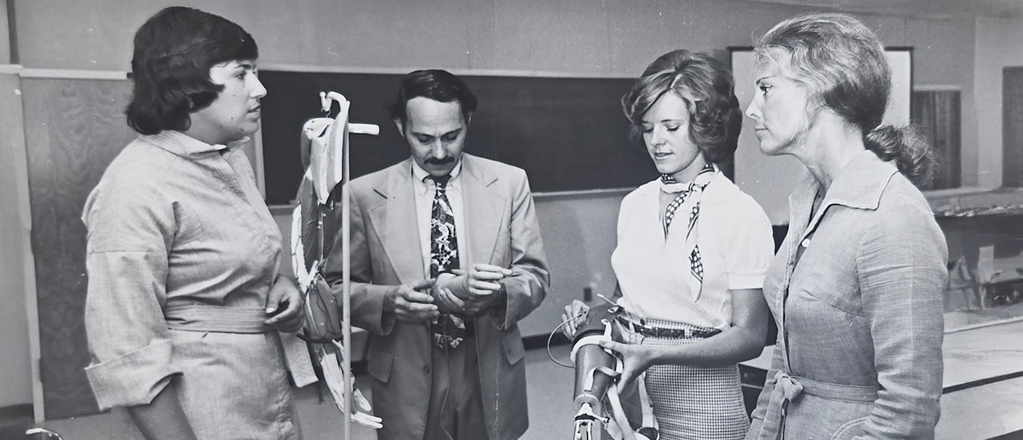 Three women and a man conversing in a classroom in the 1970s. Picture is black and white.