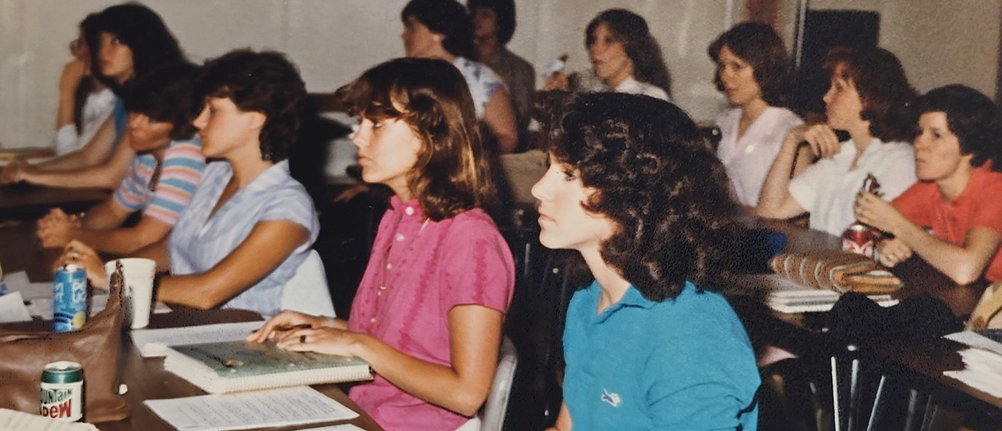 Students in a class in 1982, sitting at desks. Most are female.
