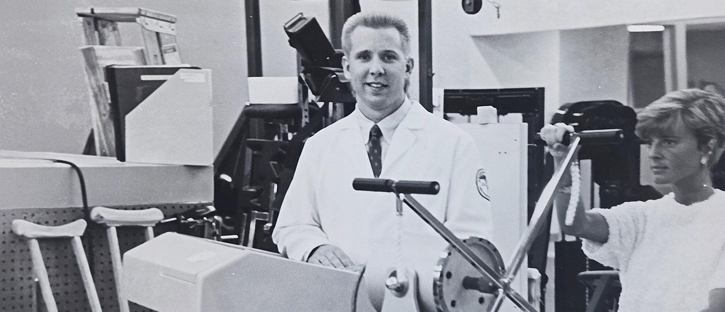 Image from 1990 of a tall man with short blond hair in a lab, wearing a white coat over shirt and tie.