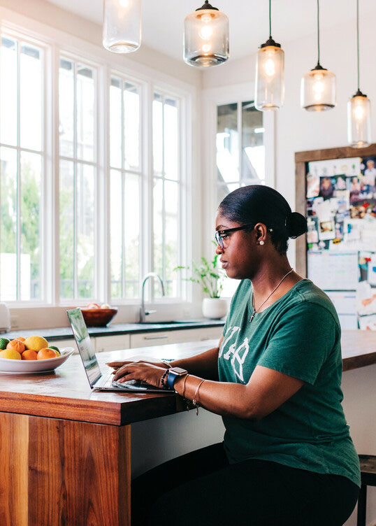 Black female working on a laptop in her home sitting at the kitchen bar.