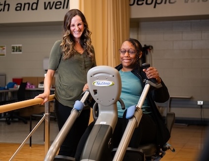 Adaptive Human Performance Lab in CEDHARS (Center for Engagement in Disability Health and Rehabilitation Sciences). Dr. Alex Evancho, PR, DPT (Assistant Professor, Physical Therapy; Director, Adaptive Human Performance Lab) is smiling while assisting a smiling staged female research participant on recumbent cross trainer machine, August 2025.